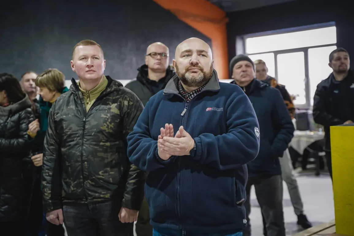 Група людей стоїть у приміщенні, одягнена в зимовий одяг — куртки та пальта. Чоловік на передньому плані в синій флісовій куртці аплодує. У фоні — вікно, стіл з речами та стіна, пофарбована в темний і помаранчевий кольори.