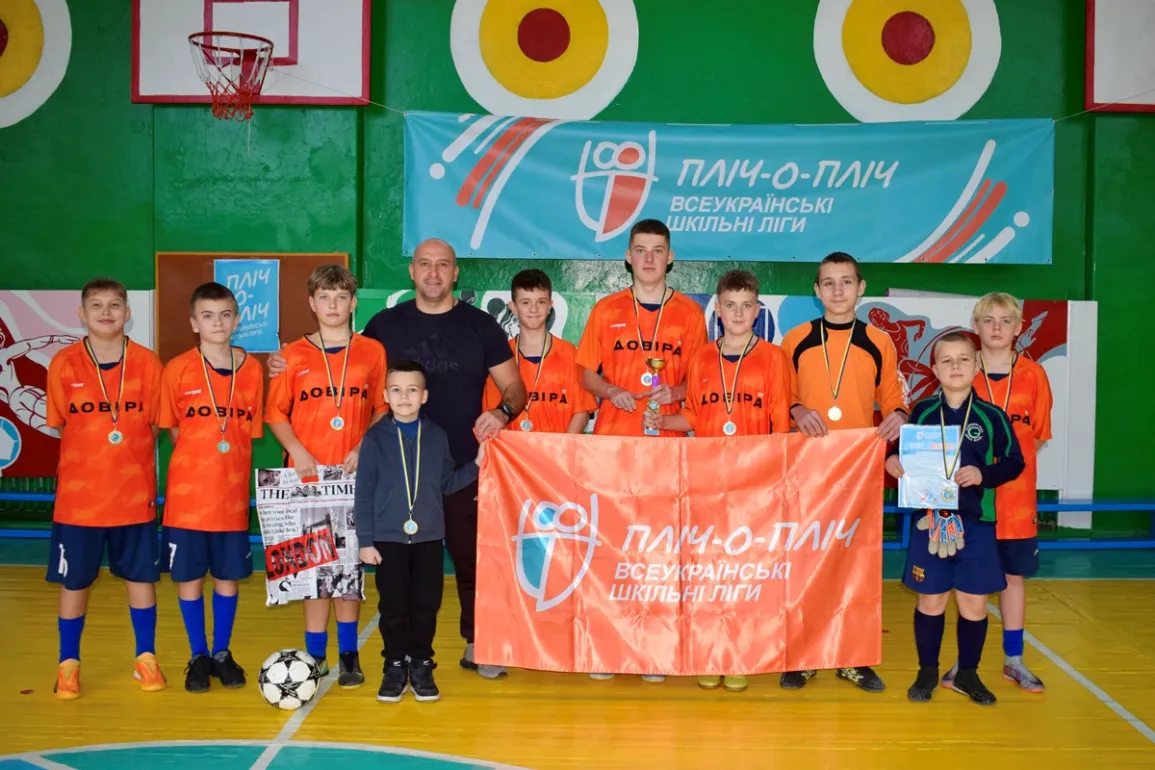 Група хлопців у спортивному залі позує для фото після змагань. Вони вдягнені в помаранчеві футболки з написом «ДОВІРА» та мають медалі на шиї. Один тримає футбольний м’яч, інший — грамоту, ще один — газету. Поруч із ними — дорослий наставник. На фоні — банер «Пліч-о-пліч. Всеукраїнські шкільні ліги», яскраві стіни та баскетбольне кільце.