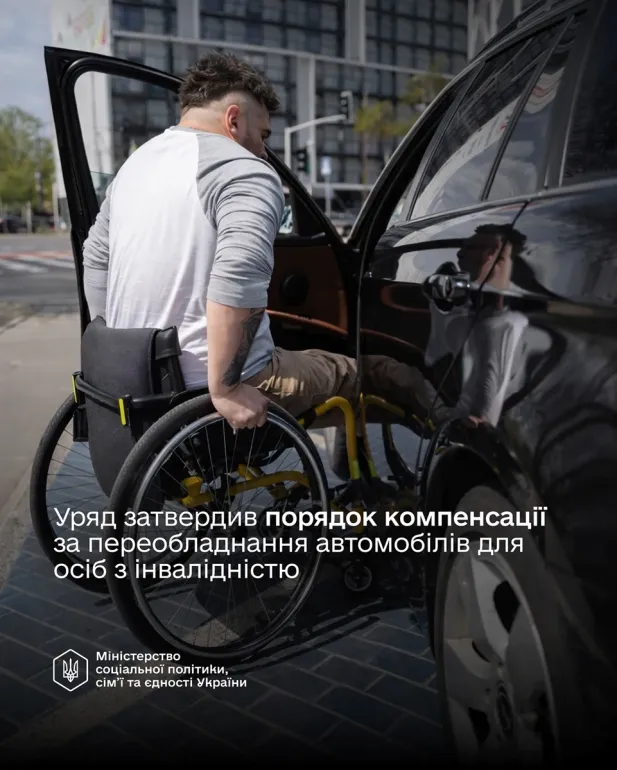 Чоловік у візку самостійно пересідає до легкового автомобіля біля сучасної будівлі та перехрестя. Він одягнений у білу футболку з сірими вставками, на лівій руці видно татуювання. Візок розташований поруч із відкритими дверима авто. На зображенні є текст українською: «Уряд затвердив порядок компенсації за переобладнання автомобілів для осіб з інвалідністю. Міністерство соціальної політики, сім’ї та єдності України».