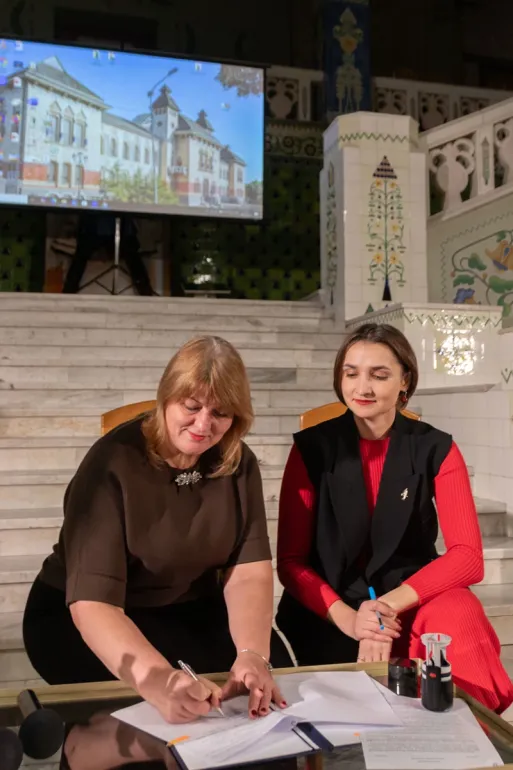 Двоє людей сидять перед сходами в урочистому приміщенні, оформленому декоративною плиткою та архітектурними елементами. Один із них підписує документ, інший уважно спостерігає, тримаючи ручку. На задньому плані — великий екран із зображенням будівлі, що, ймовірно, стосується теми заходу. Атмосфера — офіційна, з акцентом на важливість події, яка може бути частиною культурної, адміністративної або партнерської ініціативи.