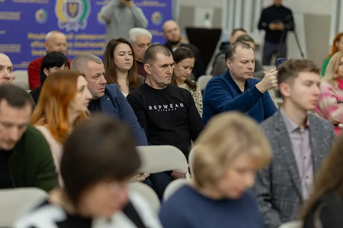 Учасники конференції сидять у залі, уважно слухаючи виступ. Один із присутніх у синьому светрі тримає смартфон, ймовірно, знімає або фотографує. На задньому плані — синій банер з гербом і кириличним текстом, що вказує на офіційний характер заходу в українськомовному регіоні.