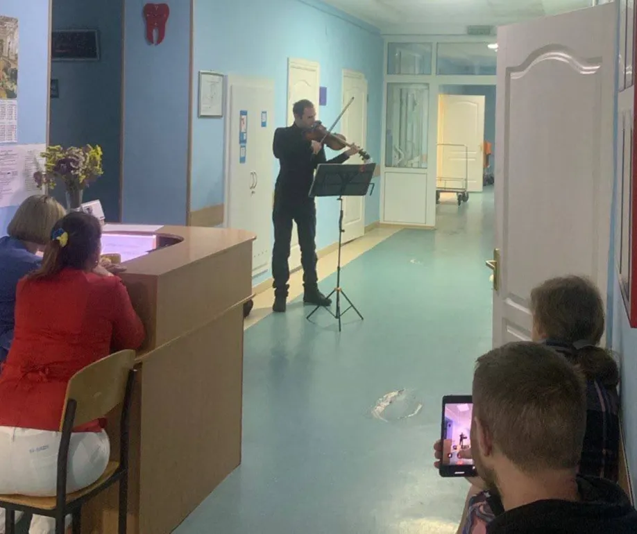 Скрипаль виконує музику у коридорі медичного закладу, стоячи перед пюпітром із нотами. Двоє жінок за рецепцією уважно слухають, а чоловік із дитиною біля дверей сидить на підлозі — чоловік знімає виступ на планшет. Яскраво освітлений простір із синіми стінами та білими дверима, на стіні — червона фігура у формі зуба, що натякає на стоматологічну тематику. Атмосфера — тепла, людяна, з акцентом на підтримку та емоційне піднесення.