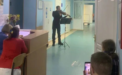 Скрипаль виконує музику у коридорі медичного закладу, стоячи перед пюпітром із нотами. Двоє жінок за рецепцією уважно слухають, а чоловік із дитиною біля дверей сидить на підлозі — чоловік знімає виступ на планшет. Яскраво освітлений простір із синіми стінами та білими дверима, на стіні — червона фігура у формі зуба, що натякає на стоматологічну тематику. Атмосфера — тепла, людяна, з акцентом на підтримку та емоційне піднесення.