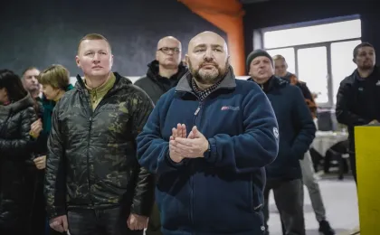 Група людей стоїть у приміщенні, одягнена в зимовий одяг — куртки та пальта. Чоловік на передньому плані в синій флісовій куртці аплодує. У фоні — вікно, стіл з речами та стіна, пофарбована в темний і помаранчевий кольори.