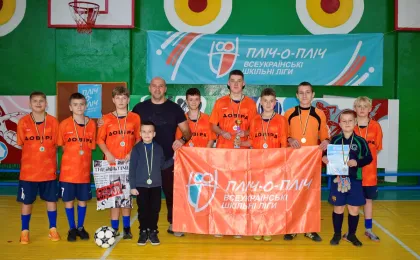 Група хлопців у спортивному залі позує для фото після змагань. Вони вдягнені в помаранчеві футболки з написом «ДОВІРА» та мають медалі на шиї. Один тримає футбольний м’яч, інший — грамоту, ще один — газету. Поруч із ними — дорослий наставник. На фоні — банер «Пліч-о-пліч. Всеукраїнські шкільні ліги», яскраві стіни та баскетбольне кільце.