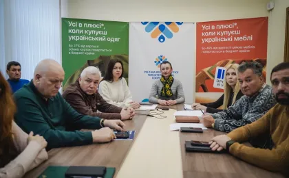 У конференц-залі за столом сидять десятеро учасників зустрічі — по п’ять з кожного боку. На задньому плані — три банери. Лівий зелений закликає купувати український одяг, адже 27% його вартості повертається в бюджет. Центральний — з логотипом Полтавського регіонального офісу міжнародного співробітництва. Правий червоний — про українські меблі, з акцентом на 46% повернення до бюджету. Атмосфера — ділова, спрямована на підтримку місцевого виробництва.