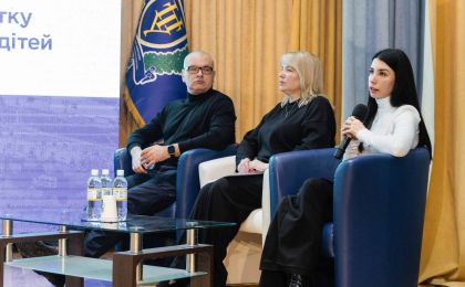 Троє учасників сидять у кріслах на сцені під час панельної дискусії. На столі перед ними — пляшки з водою та стаканчики. Один із спікерів тримає мікрофон, готуючись до виступу. На фоні — синьо-жовта завіса, прапор із гербом та екран із частковим текстом «підтримку дітей». Зображення передає атмосферу офіційного заходу, присвяченого темі захисту прав або підтримки дітей.