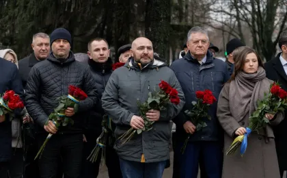 Група людей стоїть на вулиці, тримаючи букети червоних троянд, перев’язаних синьо-жовтими стрічками. Усі одягнені в зимовий одяг, на обличчях — серйозні вирази. На фоні — голі дерева, що підкреслюють холодну пору року. Зображення передає атмосферу скорботи та вшанування, ймовірно, під час пам’ятного заходу або церемонії.