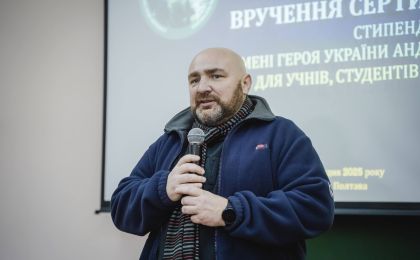 Чоловік у темно-синій куртці та смугастому шарфі виступає перед аудиторією, тримаючи мікрофон. За його спиною — екран із проєкцією, на якому українською мовою зазначено, що йдеться про церемонію вручення сертифікатів або стипендій імені Героя України Андрія. Подія присвячена учням і студентам, проходить у місті Полтава у 2025 році. Атмосфера заходу — урочиста, спрямована на вшанування пам’яті та підтримку молоді.