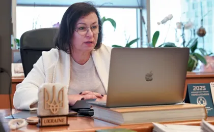 Жінка в окулярах і світлому светрі, з білим пальтом на плечах, сидить за робочим столом в офісному просторі. Вона бере участь у відеозустрічі або працює за ноутбуком Apple. На столі — велика книга, календар із написом «2025» та гаслом «Асоціація міст України — ПЕРЕМАГАЄМО», а також табличка з написом «UKRAINE» і тризубом, що підкреслює офіційний характер роботи. У приміщенні — великі вікна, кімнатні рослини та сучасні меблі. Атмосфера — професійна, з акцентом на державну або муніципальну діяльність.