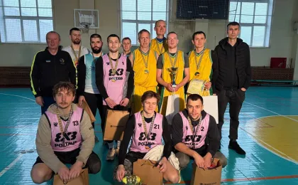 На внутрішньому баскетбольному майданчику група гравців і тренерів позує після завершення змагань. Команди одягнені у жовті форми та рожеві манішки поверх темного одягу. Учасники тримають кубки, медалі, грамоти та подарункові пакети з логотипом «FERREXPO», що свідчить про церемонію нагородження. На задньому плані — вимкнене табло та великі вікна, крізь які проникає природне світло.