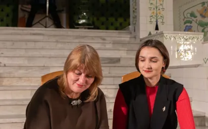 Двоє людей сидять перед сходами в урочистому приміщенні, оформленому декоративною плиткою та архітектурними елементами. Один із них підписує документ, інший уважно спостерігає, тримаючи ручку. На задньому плані — великий екран із зображенням будівлі, що, ймовірно, стосується теми заходу. Атмосфера — офіційна, з акцентом на важливість події, яка може бути частиною культурної, адміністративної або партнерської ініціативи.