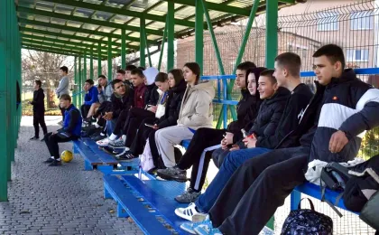 На зображенні — кілька юнаків і дівчат у спортивному одязі та куртках сидять на синіх металевих лавках під зеленою металевою конструкцією. Поряд — жовтий футбольний м’яч, а за трибунами видно огорожу, що вказує на спортивний майданчик або шкільне подвір’я. Дехто спілкується, інші просто очікують — можливо, на початок гри чи заходу. Атмосфера — невимушена, з елементами очікування та товариського спілкування.