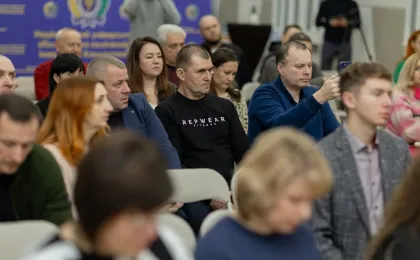 Учасники конференції сидять у залі, уважно слухаючи виступ. Один із присутніх у синьому светрі тримає смартфон, ймовірно, знімає або фотографує. На задньому плані — синій банер з гербом і кириличним текстом, що вказує на офіційний характер заходу в українськомовному регіоні.