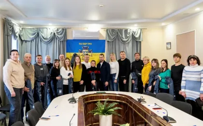 У конференц-залі група людей позує для спільного фото на тлі банера з написом "116 ОБР ТРО" та гербом, що символізує 116-ту окрему бригаду територіальної оборони України. Учасники тримають у руках форму або спорядження, що свідчить про офіційний захід — можливо, вручення екіпірування чи зустріч з представниками бригади. На столі перед ними — мікрофони, ручки та блокноти, що підкреслює формальність події. Атмосфера знімка передає дух організованості, підтримки та спільної участі у справі національної безпеки