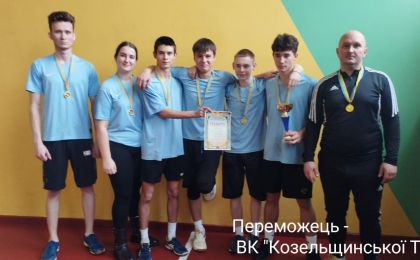 Група з семи спортсменів позує перед жовто-зеленою стіною після перемоги в змаганнях. Шестеро з них одягнені в блакитні спортивні футболки та чорні штани, один — у чорному спортивному костюмі з білими смужками. Усі мають золоті медалі на шиї. Один учасник тримає грамоту, інший — трофей. На стіні напис українською: «Переможець — ВК 'Козельщинської ТГ'».