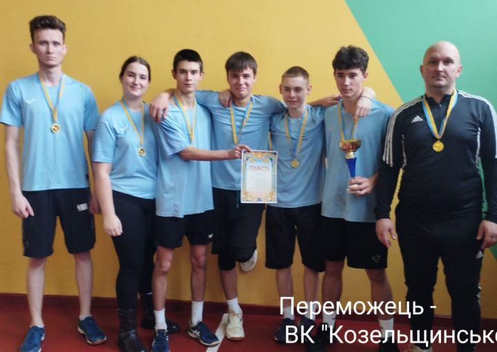 Група з семи спортсменів позує перед жовто-зеленою стіною після перемоги в змаганнях. Шестеро з них одягнені в блакитні спортивні футболки та чорні штани, один — у чорному спортивному костюмі з білими смужками. Усі мають золоті медалі на шиї. Один учасник тримає грамоту, інший — трофей. На стіні напис українською: «Переможець — ВК 'Козельщинської ТГ'».